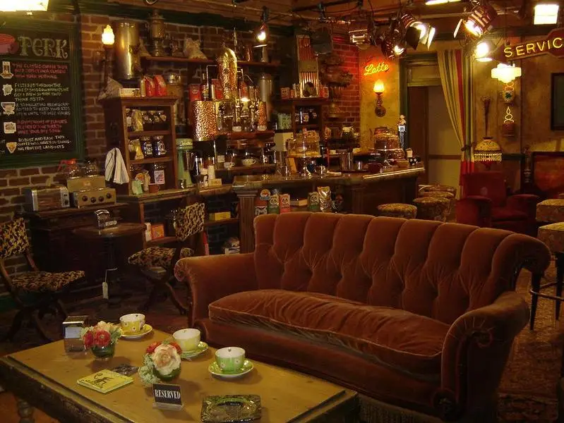 O icônico sofá laranja aveludado do Central Perk em primeiro plano, com o balcão de café e a parede de tijolos cheia de equipamentos de café e objetos de decoração ao fundo.