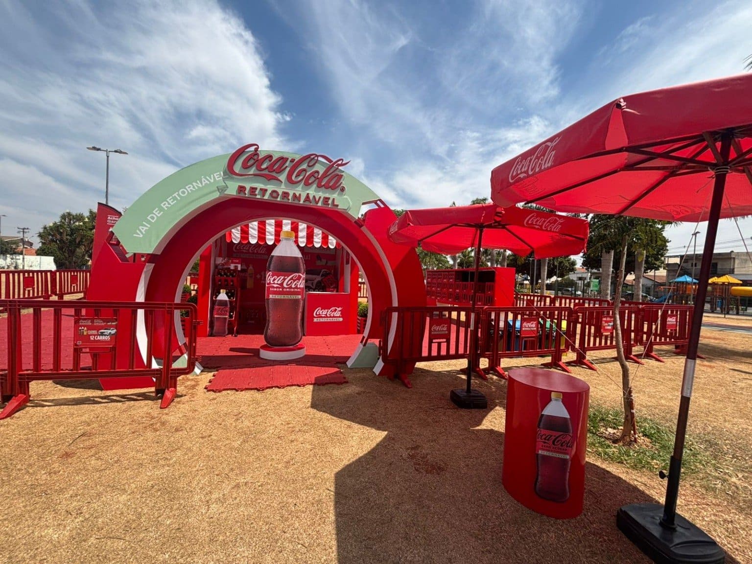 Entrada de evento da Coca-Cola com arco vermelho, garrafa gigante e guarda-sóis, promovendo o tema "Coca-Cola Retornável".