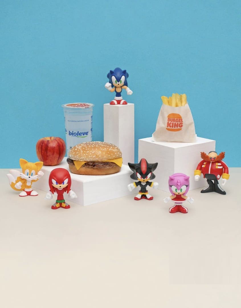 Brinquedos da coleção Sonic no Burger King junto a itens do Combo King Jr.: hambúrguer, batatas fritas, iogurte e maçã.