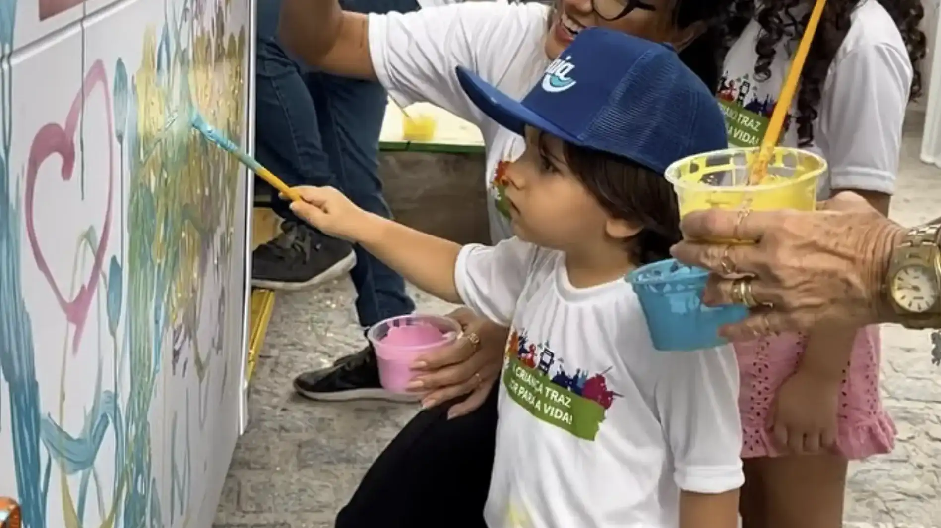 Uma criança com boné azul e camiseta branca pintando a lateral de um ônibus com um pincel.