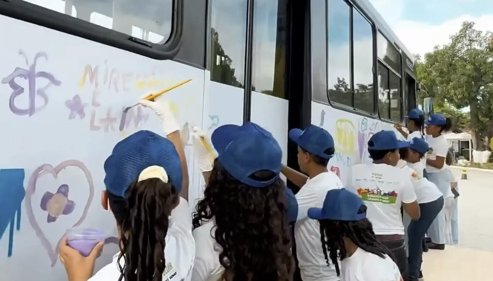 Crianças pintando a lateral de um ônibus branco com pincéis.