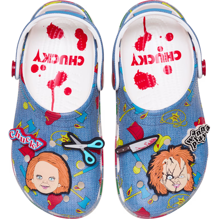 Visão superior de um par de Crocs temáticos do Chucky. A estampa imita jeans com desenhos de objetos do filme. As palmilhas brancas têm respingos de sangue e a palavra "CHUCKY". Os Jibbitz incluem faca, tesoura e duas versões da cabeça do boneco.