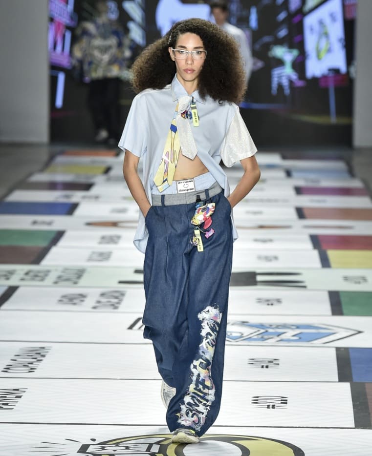 Modelo com cabelo cacheado volumoso desfilando na passarela da SPFW, usando uma camisa azul-clara cropped com gravata e calça jeans larga com estampa.