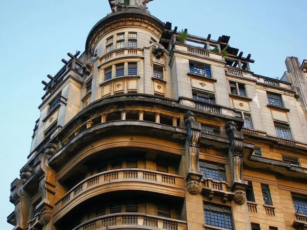 Vista angular do Edifício Rolim, mostrando sua fachada curva em pedra clara, balcões com balaustradas e detalhes arquitetônicos ornamentados sob um céu azul claro.