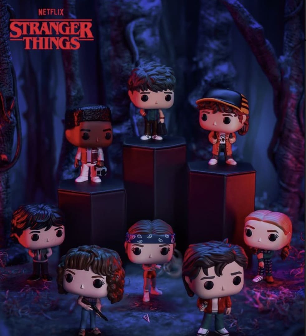 Imagem promocional de sete figuras Funko Pop! de personagens de Stranger Things, incluindo Lucas, Dustin, Mike, Eleven, Max e outros. Os bonecos estão dispostos em diferentes níveis em um cenário escuro que r