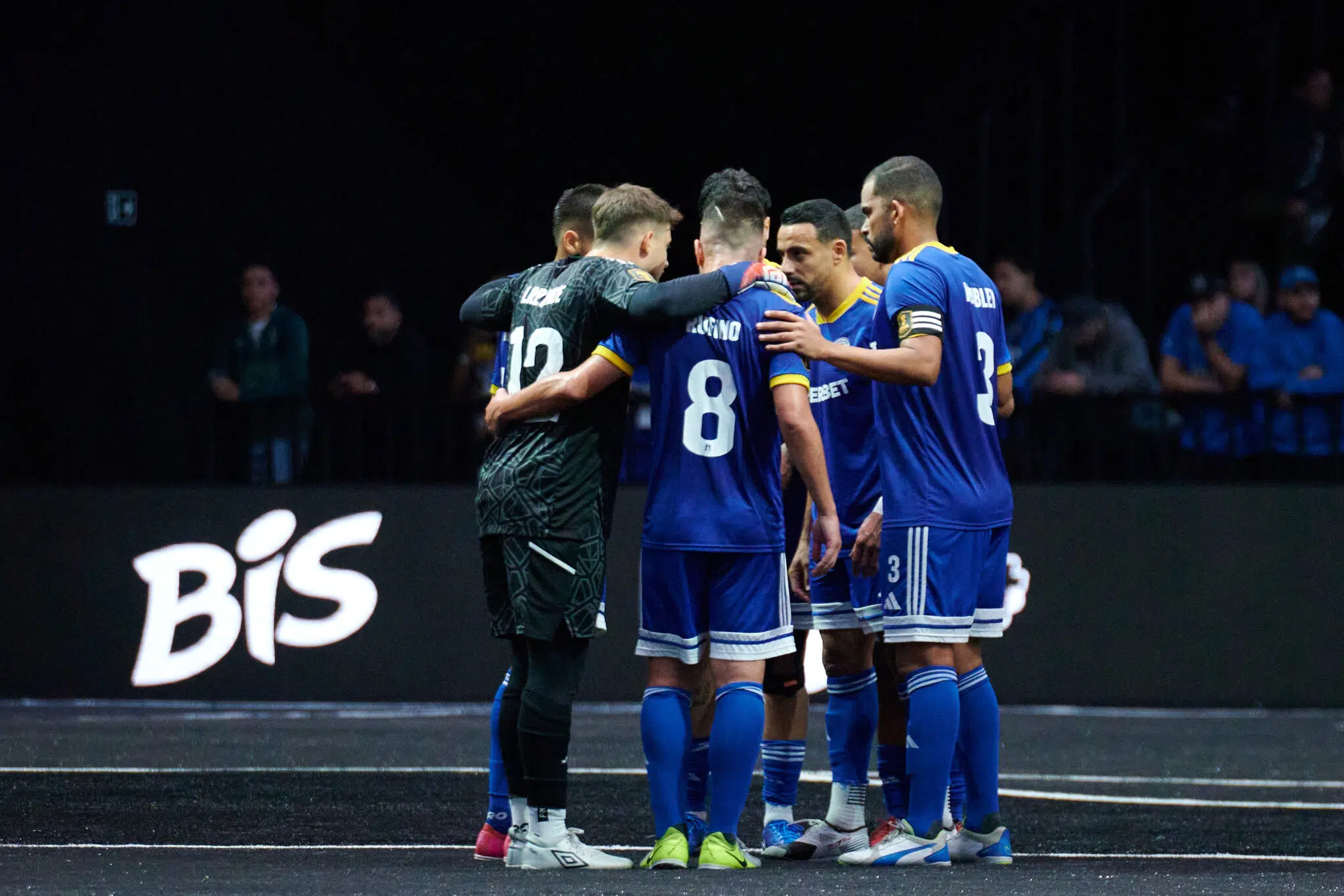 Time de futebol com uniformes azuis e pretos reunido em círculo em campo de jogo, com a logo "Bis" em destaque no chão.