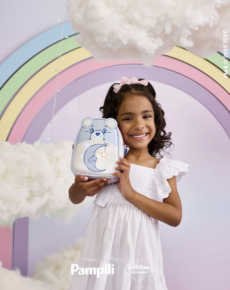 Menina sorrindo, segurando uma pequena bolsa azul em formato de urso da coleção Pampili Ursinhos Carinhosos, em um cenário de nuvens e arco-íris pastel.