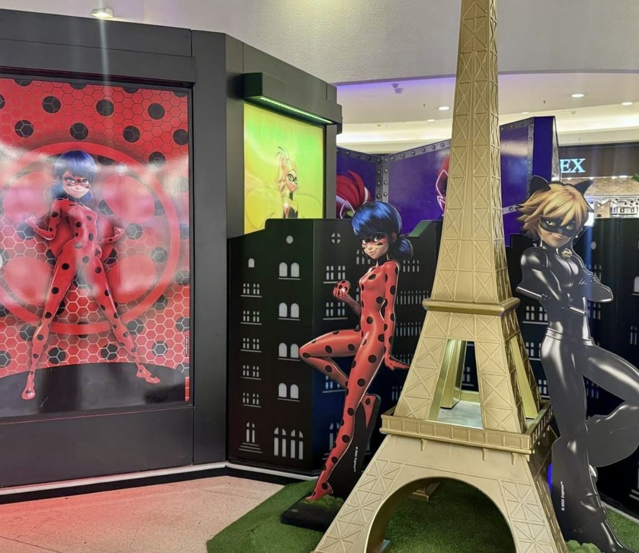 Área de fotos de Miraculous com um modelo dourado da Torre Eiffel e recortes de papelão de Ladybug e Cat Noir.