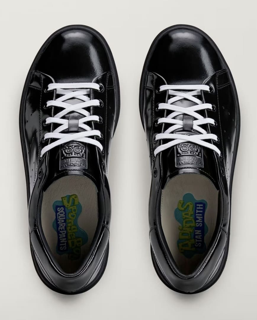 Visão superior de um par de tênis Adidas Stan Smith Freizeit preto com cadarços brancos, destacando as palmilhas personalizadas com a marca "SpongeBob SquarePants" no pé esquerdo e "Adidas Stan Smith" no pé direito.