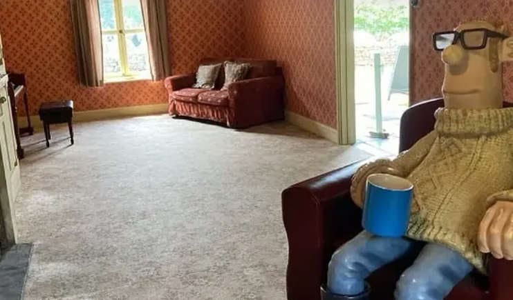 A imagem mostra um cômodo com papel de parede avermelhado e um tapete claro. No canto direito, uma estátua do Fazendeiro de Shaun, o Carneiro, está sentada em uma poltrona vermelha, segurando uma caneca azul. Ao fundo, um sofá vermelho e uma janela iluminada.