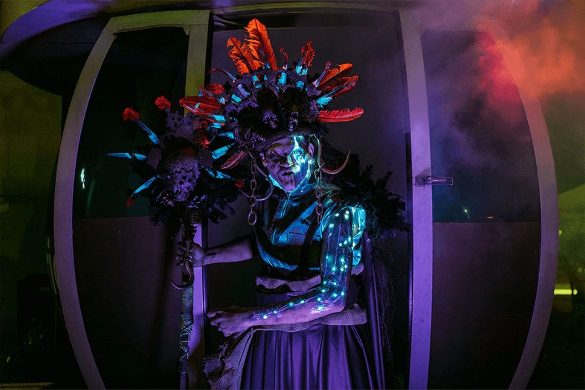 Ator com fantasia de criatura xamânica ou tribal com cocar de penas laranjas e detalhes luminosos azuis, emergindo de uma cabine da Roda Rico em São Paulo sob forte iluminação roxa e escura.