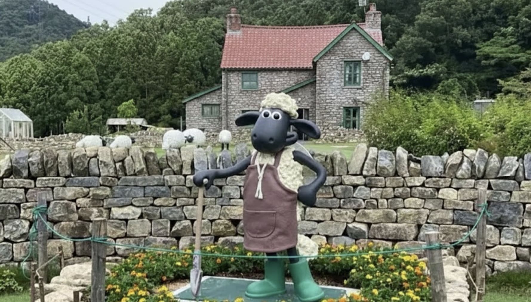 Estátua do Shaun, o Carneiro, em pé, segurando uma pá, em frente a um muro de pedra e uma casa de fazenda, com árvores e colinas verdes ao fundo.