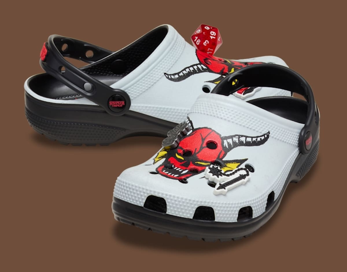 Um par de Crocs brancos e pretos da coleção Stranger Things com a estampa da cabeça do Demogorgon e um dado vermelho Jibbitz.