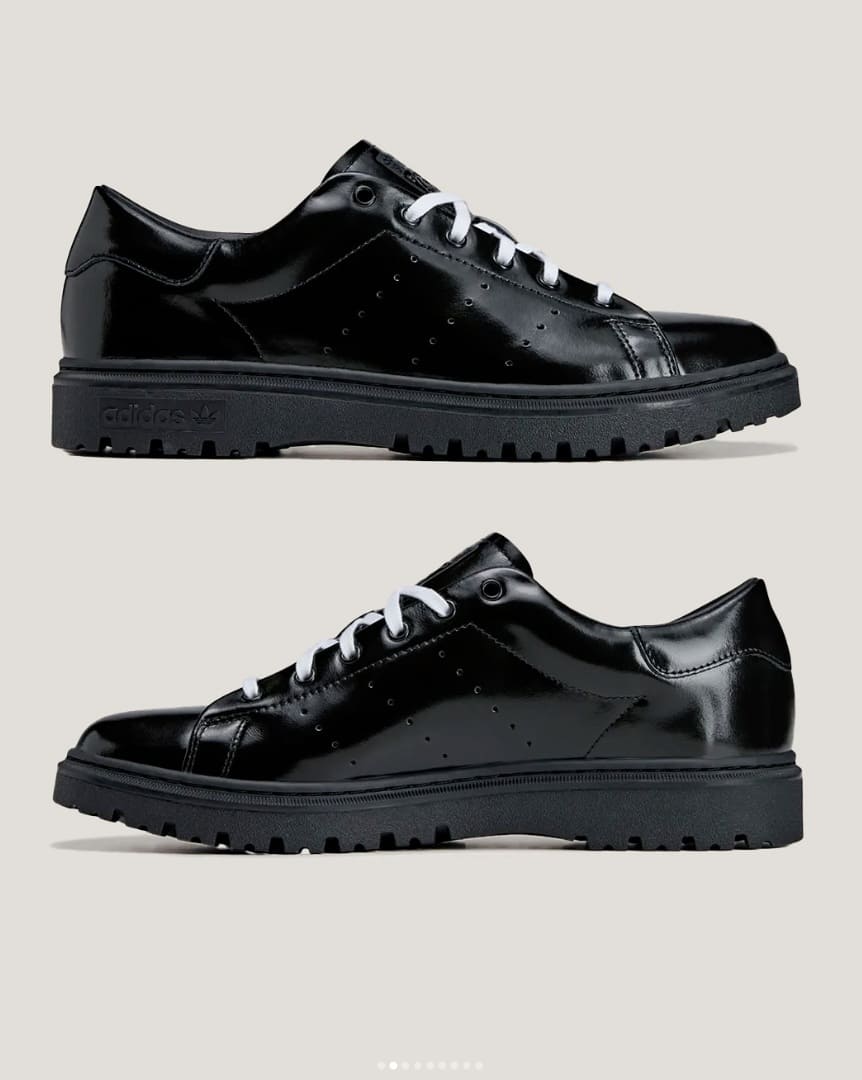 Dois tênis Adidas Stan Smith Freizeit pretos brilhantes com cadarços brancos, dispostos um acima do outro e mostrando o perfil lateral, destacando o solado tratorado.