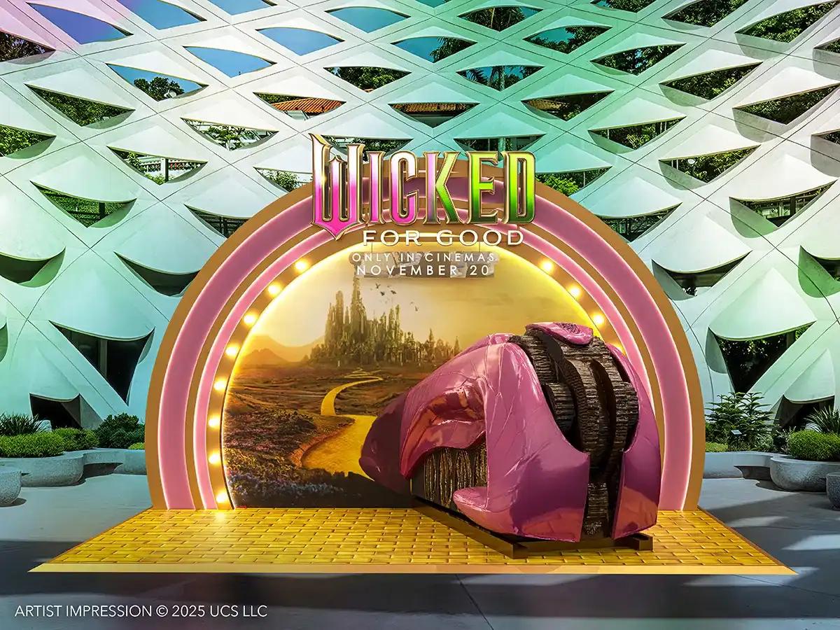 Instalação promocional externa do filme "Wicked For Good" com a logo e uma representação da Estrada de Tijolos Amarelos e Cidade Esmeralda, com as Pernas da Bruxa caídas.