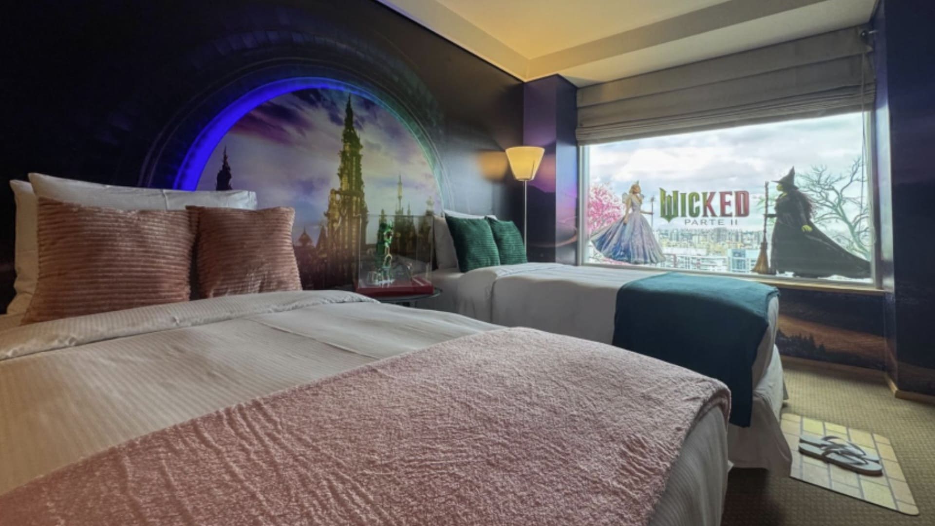 Quarto de hotel temático de Wicked, com duas camas, almofadas rosa e verde, um mural na parede com um arco e a cidade de Oz ao fundo, e um adesivo grande na janela com a logo "Wicked Parte II" e as duas bruxas.