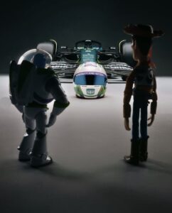 Os bonecos de Buzz Lightyear e Woody estão de costas, olhando para um capacete de piloto customizado de Buzz e, ao fundo, para um carro de Fórmula 1 da Aston Martin.