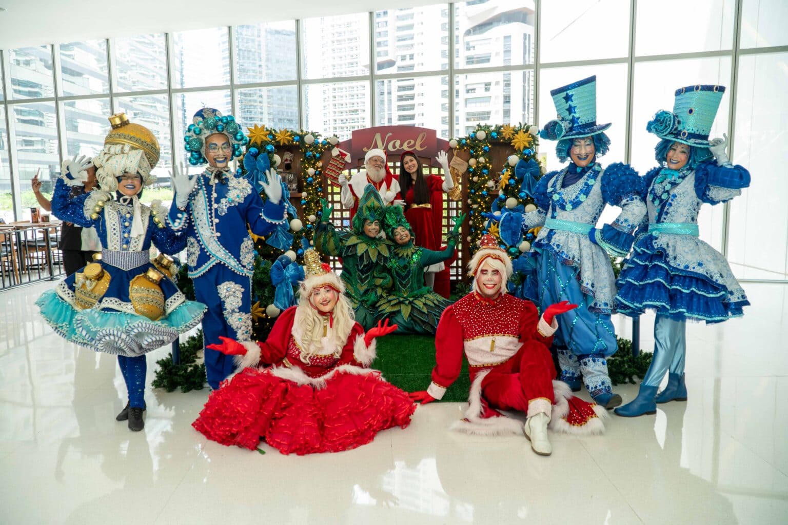 Grupo de artistas em trajes de Natal coloridos (vermelho, azul e verde) posando no cenário "Noel" de um shopping center.