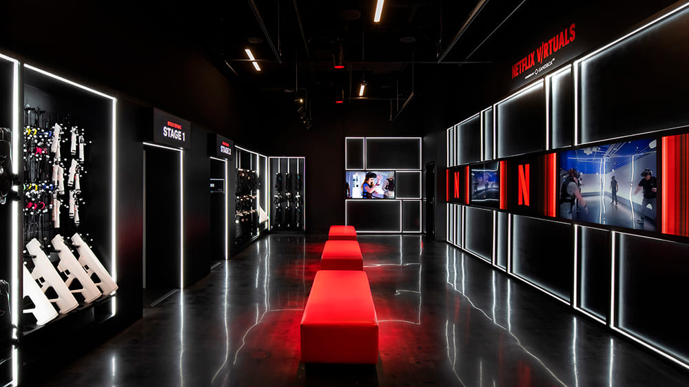 Interior escuro e moderno da "Netflix Virtually" com bancos longos e vermelhos no centro, paredes pretas e luzes brancas de neon que contornam painéis e telas. Equipamentos de jogo (armas e coletes) estão dispostos em expositores nas laterais.