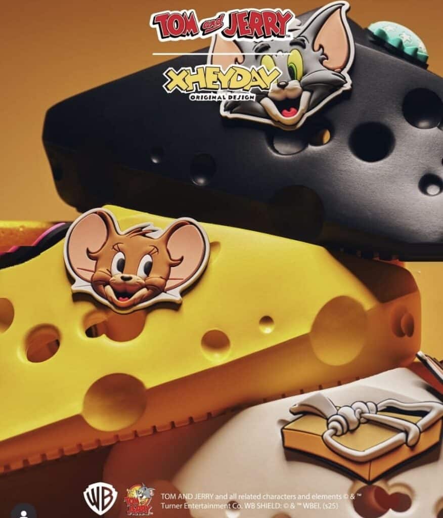 Close-up de três chinelos slip-on Tom e Jerry da XHeyDay: um preto com um pin do Tom, um amarelo com um pin do Jerry e um branco com um pin de um presente.
