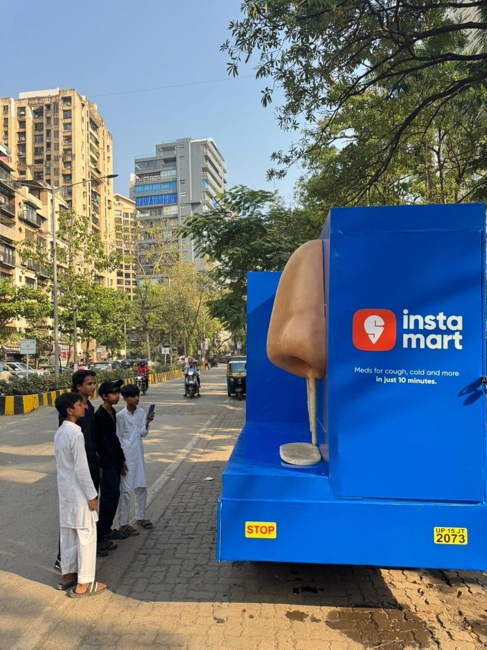 Campanha de marketing Out-of-Home (OOH) da Instamart: a lateral azul de um caminhão em uma rua urbana exibe um nariz humano gigante em 3D e o logotipo da Instamart com a mensagem "Meds for cough, cold and more in just 10 minutes." Um pequeno grupo de pessoas está olhando para a instalação.