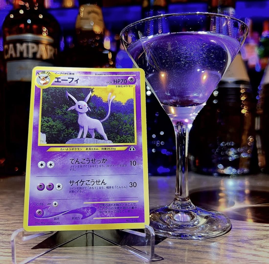 Coquetel roxo em taça de Martini ao lado de uma carta de Pokémon do Espeon.