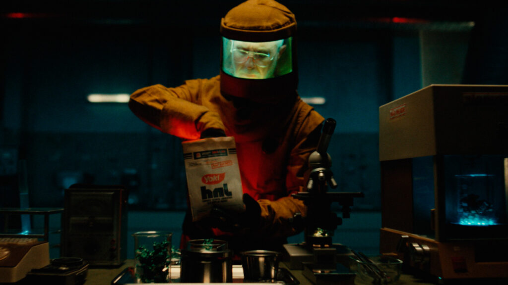 Homem de óculos, vestindo um traje de proteção laranja, semelhante a um Hazmat, em um laboratório escuro e mal iluminado, segurando um pacote de pipoca Yoki Huk. Um microscópio e equipamentos científicos com luz azul e verde estão sobre a mesa.
