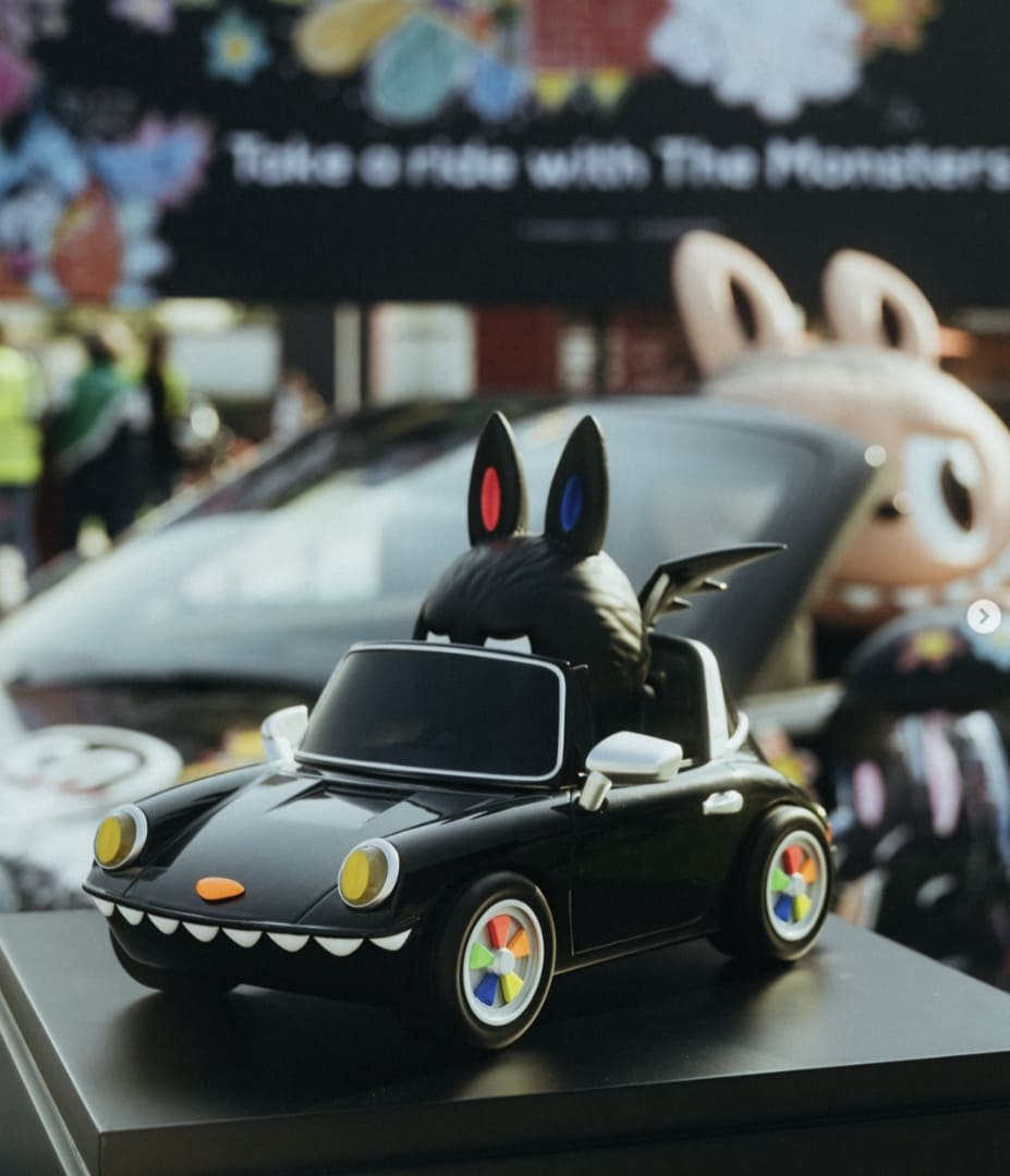 Miniatura de carro conversível preto com estilo de brinquedo, rodas coloridas, boca de dentes pontudos e um boneco Labubu preto sentado. Ao fundo, um carro Porsche e um boneco Labubu maior.