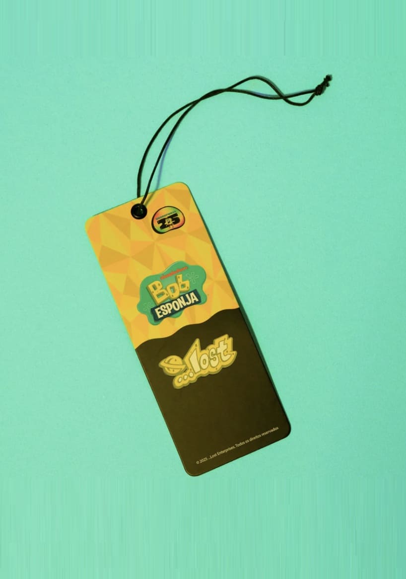 Etiqueta (tag) retangular da colaboração Lost e Bob Esponja, com a parte superior amarela e verde apresentando o logo do Bob Esponja e a parte inferior preta com o logo da Lost, suspensa por um cordão preto em um fundo azul-claro.