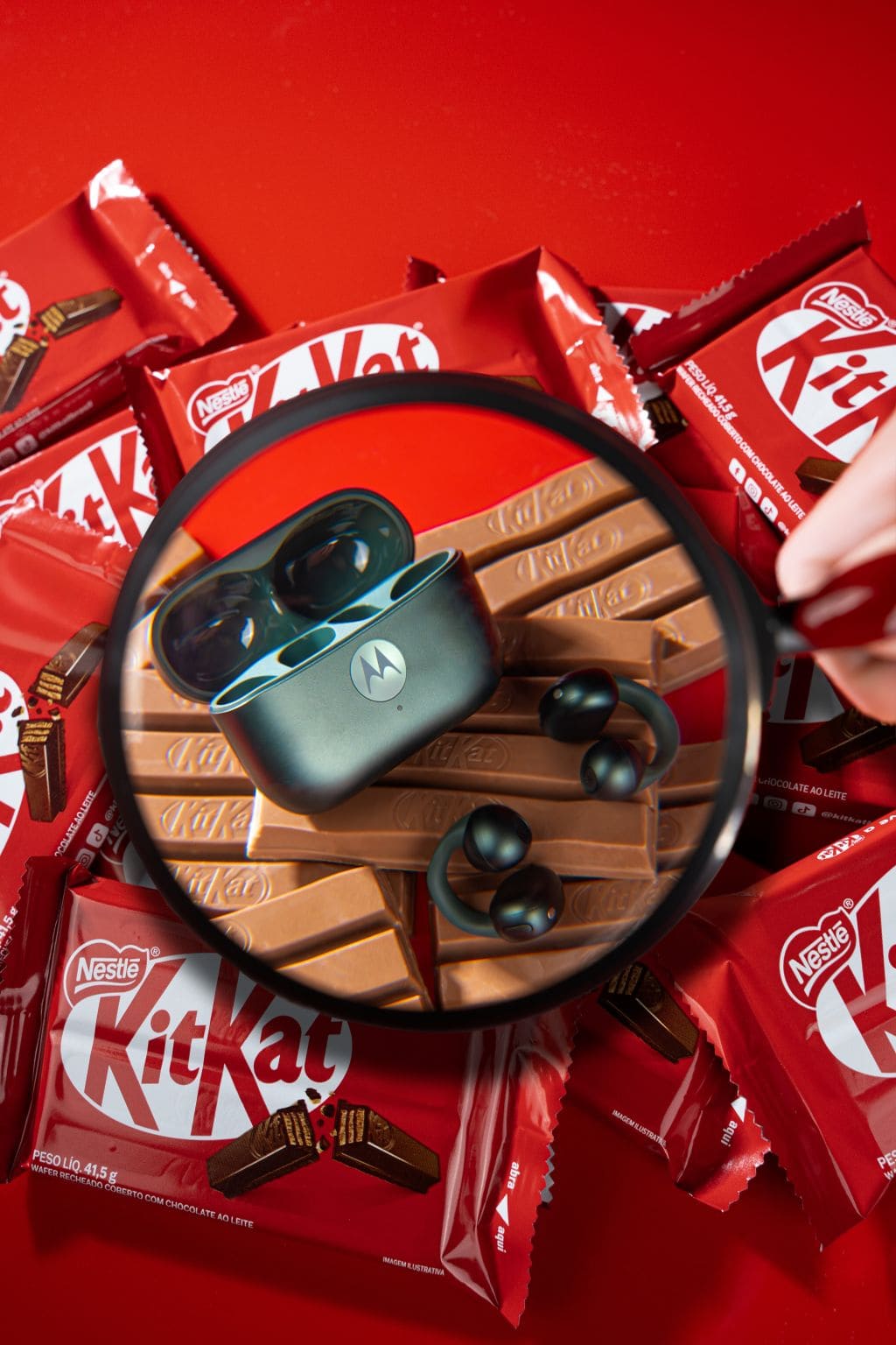 Estojo de fones de ouvido Motorola na cor cinza-chumbo e fones pretos com design de gancho, vistos através de uma lupa sobre barras de chocolate KitKat.