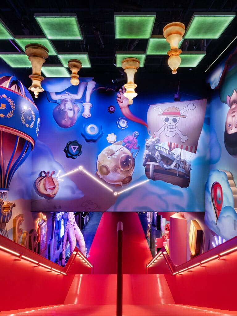 Escadaria vermelha vibrante e iluminada por neon que leva a um salão imersivo no teto. O teto apresenta um mural que combina elementos de diferentes séries da Netflix, incluindo peças de xadrez gigantescas penduradas (referência a O Gambito da Rainha), o Jolly Roger de One Piece e outros ícones flutuantes contra um céu azul.