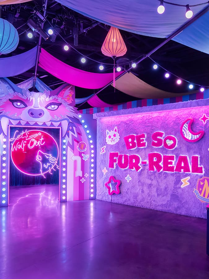 Sala de ativação temática da série Wolf Like Me na Netflix House, decorada em tons vibrantes de rosa e roxo neon. A entrada principal tem o formato da cabeça de um lobo e um sinal de neon que diz "Let's Wolf Out". A parede lateral é forrada com material peludo e tem o letreiro "BE SO FOR-REAL".