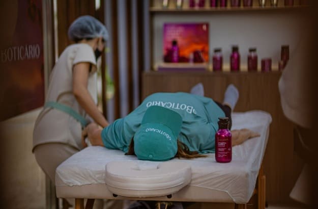 Pessoa deitada de bruços em uma maca de massagem, com boné e camiseta d'O Boticário, recebendo uma massagem de uma terapeuta. Produtos da marca estão visíveis ao fundo.