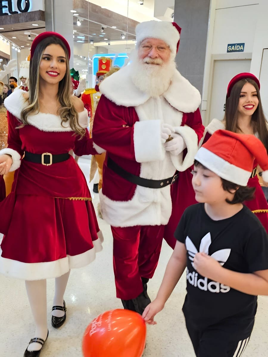 Papai Noel caminhando em um shopping, ladeado por duas ajudantes vestidas de Mamãe Noel e uma criança com gorro.