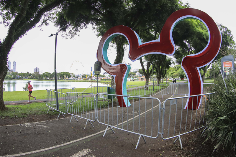 Arco de orelhas do Mickey em um parque perto de um lago, com uma pessoa correndo e uma roda-gigante ao fundo.