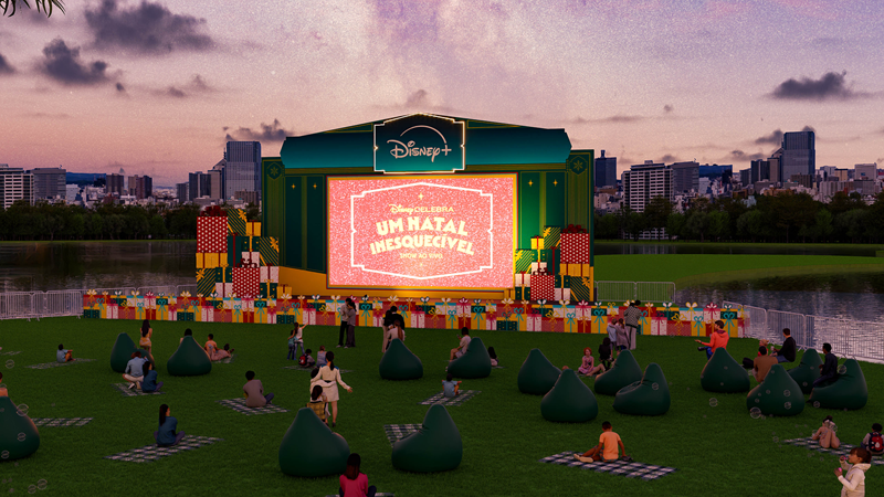 Representação de um cinema ou evento da Disney+ ao ar livre, com um palco verde, uma tela grande exibindo "Um Natal Inesquecível", pessoas sentadas em pufes verdes e mantas na grama, em frente a um lago e o skyline da cidade ao entardecer.
