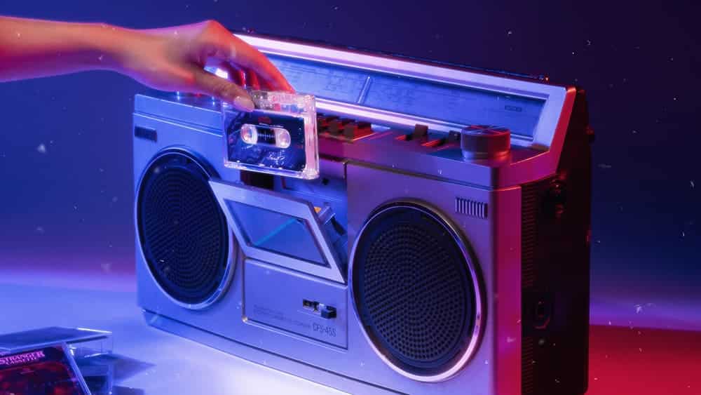Uma mão inserindo uma fita cassete transparente em um boombox prateado retrô, sob iluminação neon azul e rosa.