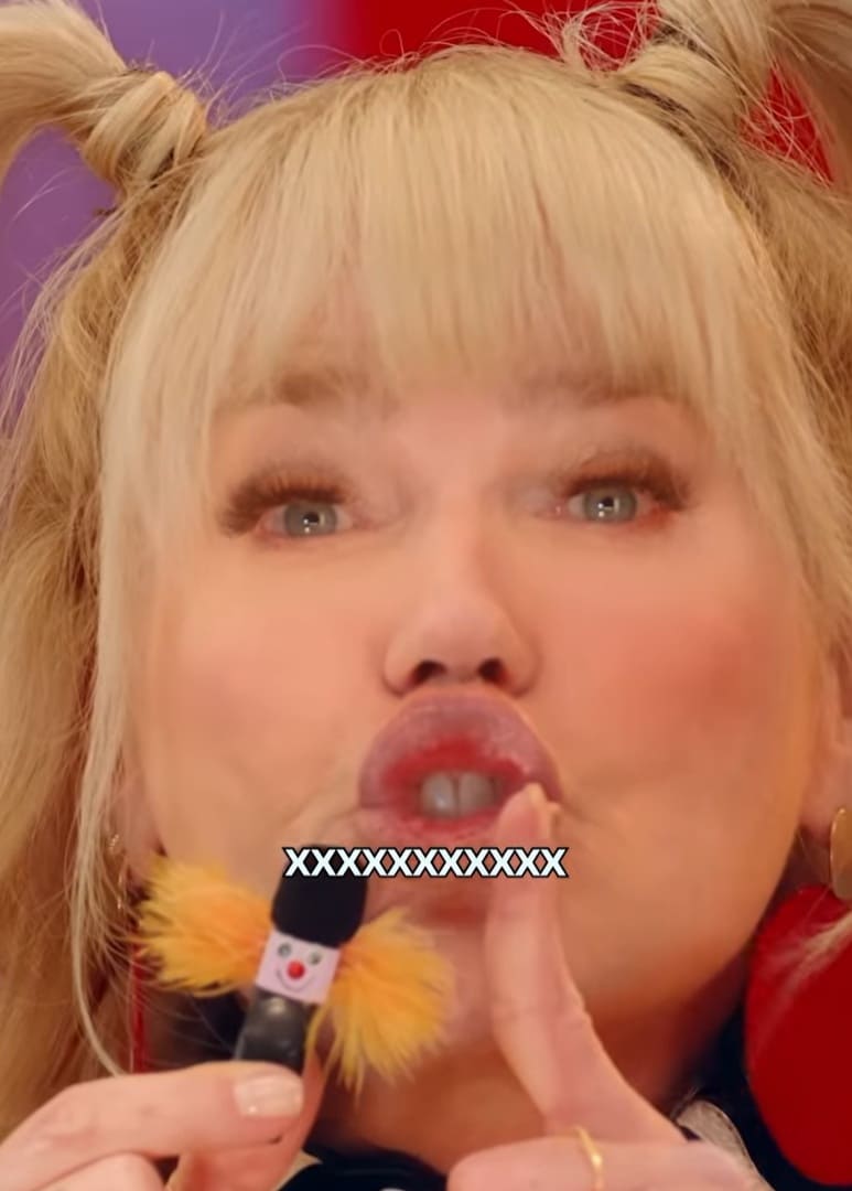 Xuxa, com cabelo loiro em coques e franja, faz bico com os lábios enquanto segura um pequeno microfone decorado com pompons amarelos e uma carinha. Seu dedo indicador está sobre os lábios, pedindo silêncio. Ela usa brincos grandes e dourados.