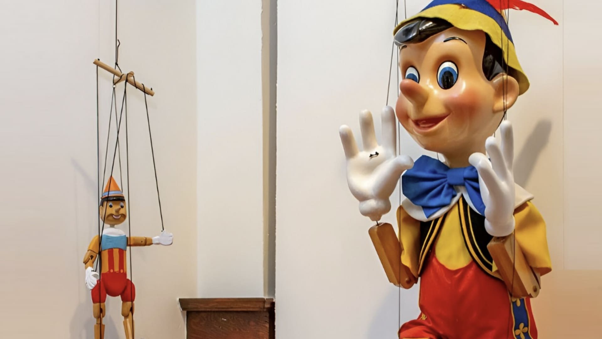 Dois bonecos de Pinóquio suspensos por cordas em uma exposição. Um é grande e moderno, e o outro é menor e tradicional.