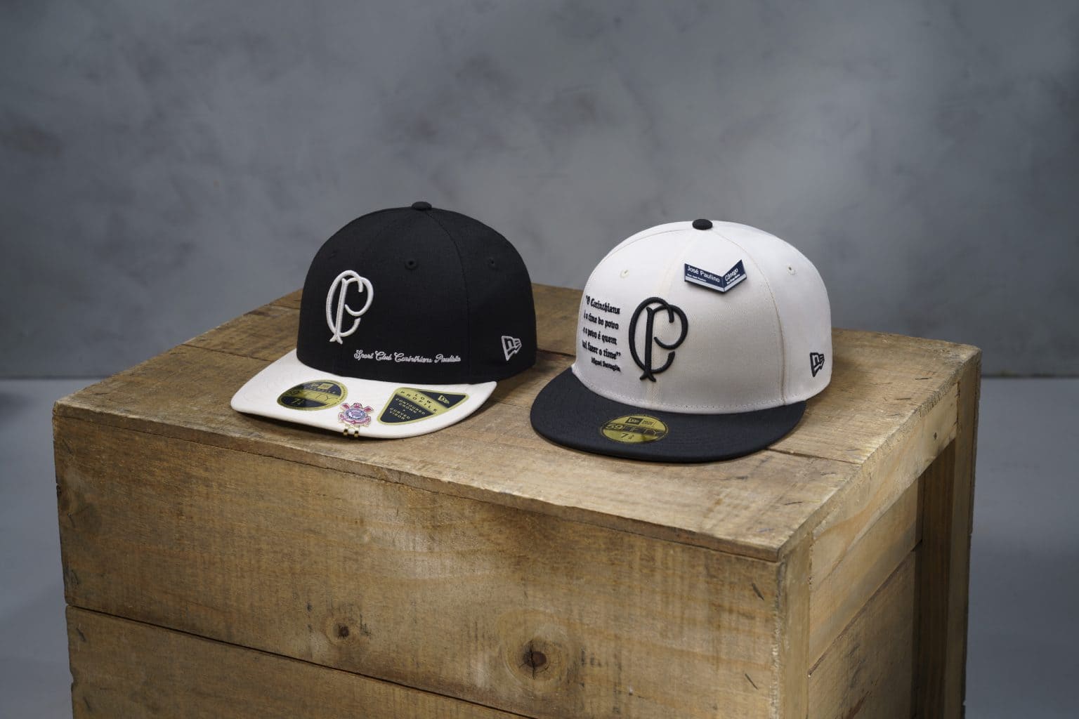 Dois bonés da coleção New Era x Corinthians sobre um caixote de madeira. Um modelo é preto com aba branca e o outro é branco com aba preta, ambos exibindo o escudo retrô "CP" e detalhes históricos.