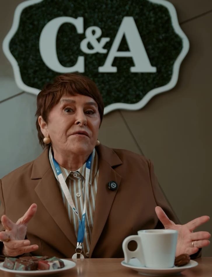 Márcia Sensitiva sentada em uma mesa, em frente ao logo da C&A, em um ambiente interno.