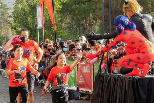 Crianças sorrindo e acenando em uma corrida temática, com pessoas fantasiadas de Ladybug e Cat Noir em um palco ao lado.