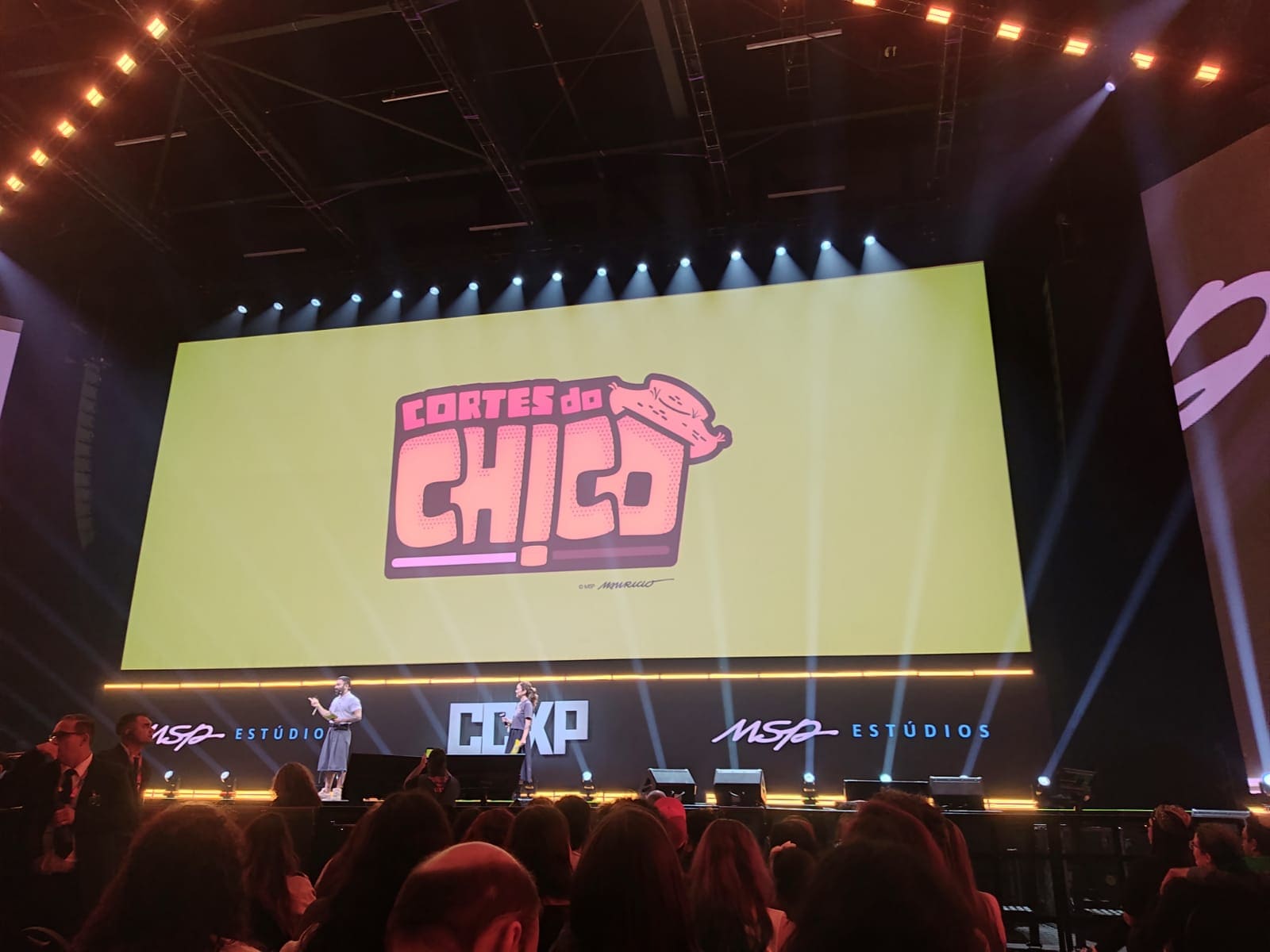 Banner amarelo grande em um palco de evento com o título "CORTES DO CHICO" em fonte estilizada, na frente de um bonezinho do Chico Bento, com a marca "MSP ESTÚDIOS CCXP" abaixo. Duas pessoas estão no palco em frente ao banner, falando para uma plateia.