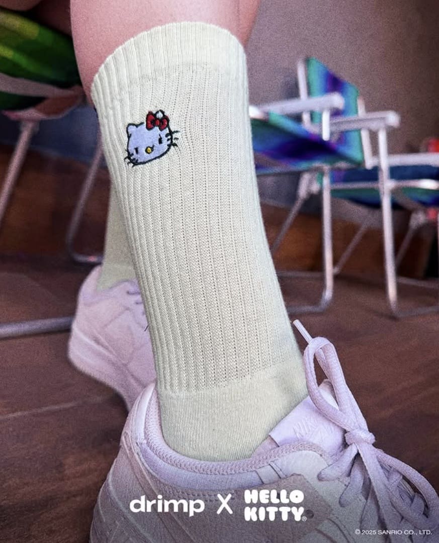 Close-up da perna de uma pessoa usando uma meia de cano alto creme canelada com bordado da Hello Kitty e tênis branco. O logo "drimp X HELLO KITTY" está sobreposto na parte inferior.