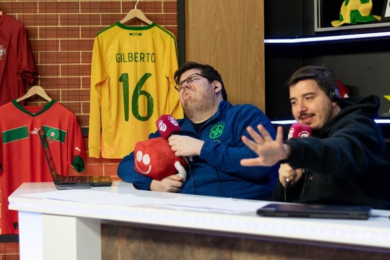 Duas pessoas, sentadas em uma mesa de estúdio de transmissão esportiva, interagem com microfones e um boneco vermelho. Camisas de seleções de futebol, incluindo uma do Brasil com o nome 'GILBERTO 16', estão penduradas ao fundo.