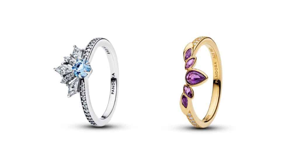 Dois anéis da Pandora com fundo branco: o da esquerda é prateado com uma pedra azul central e cristais ao redor em formato de floco de neve (representando Elsa); o da direita é dourado com pedras roxas em formato de folha/gota (representando Anna).