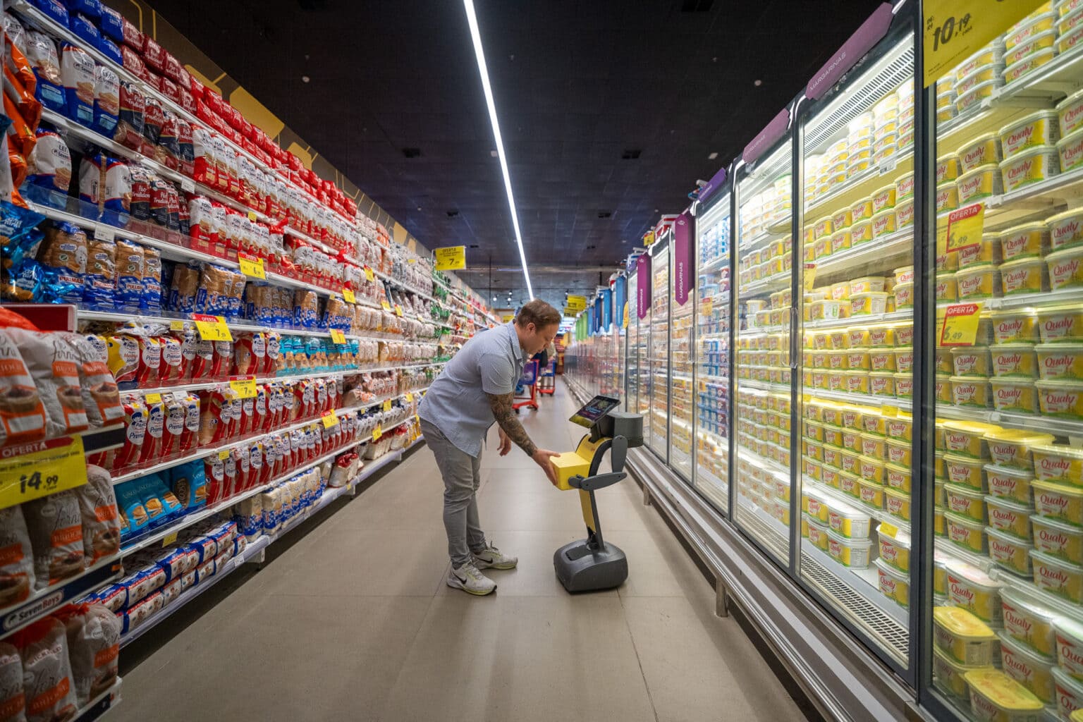 Um robô de publicidade OOH (Out-of-Home), amarelo e cinza, interagindo com um consumidor em um corredor de supermercado.
