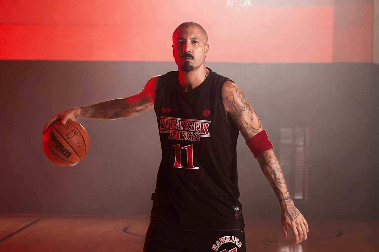 Homem careca e tatuado, vestindo uma regata de basquete preta com "STRANGER THINGS 11" estampado, segurando uma bola de basquete Wilson em uma quadra com luz vermelha.